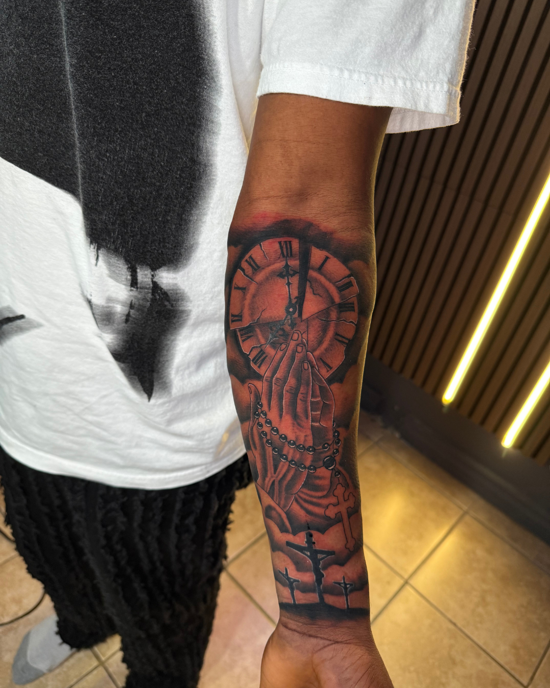 Black and grey praying hands with a rosary, clock, and crosses on the forearm