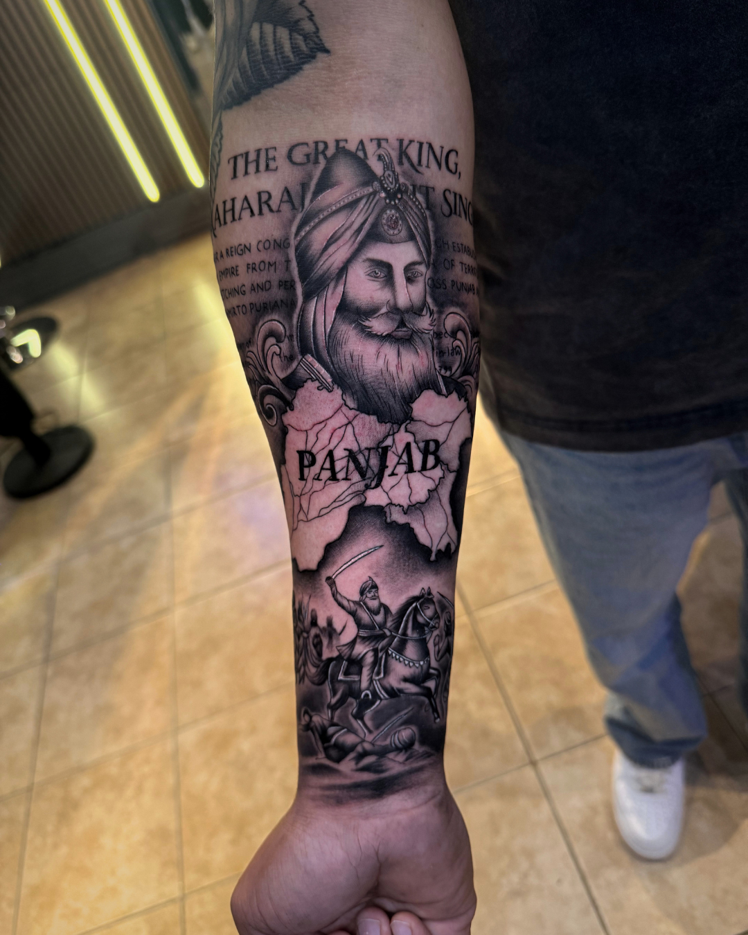 Black and grey portrait of Maharaja Ranjit Singh with a map of Panjab and a battle scene on the forearm