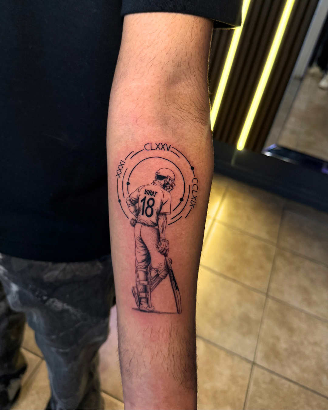 Detailed black and grey tattoo of Virat Kohli with a bat, roman numerals, and geometric elements on the forearm
