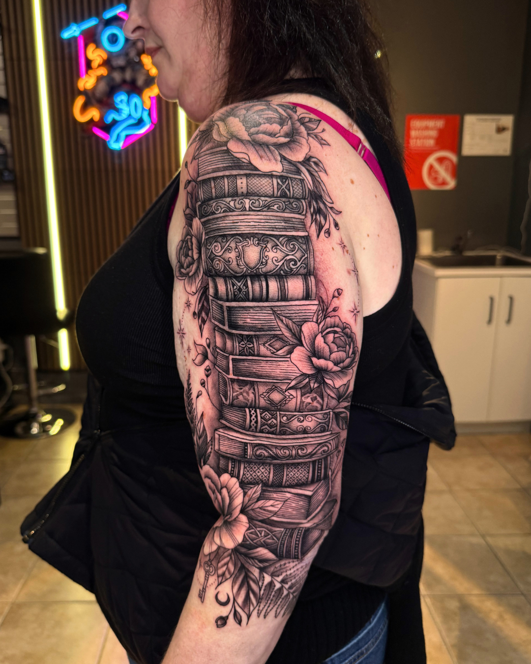 Black and grey tattoo of stacked vintage books surrounded by flowers on the arm