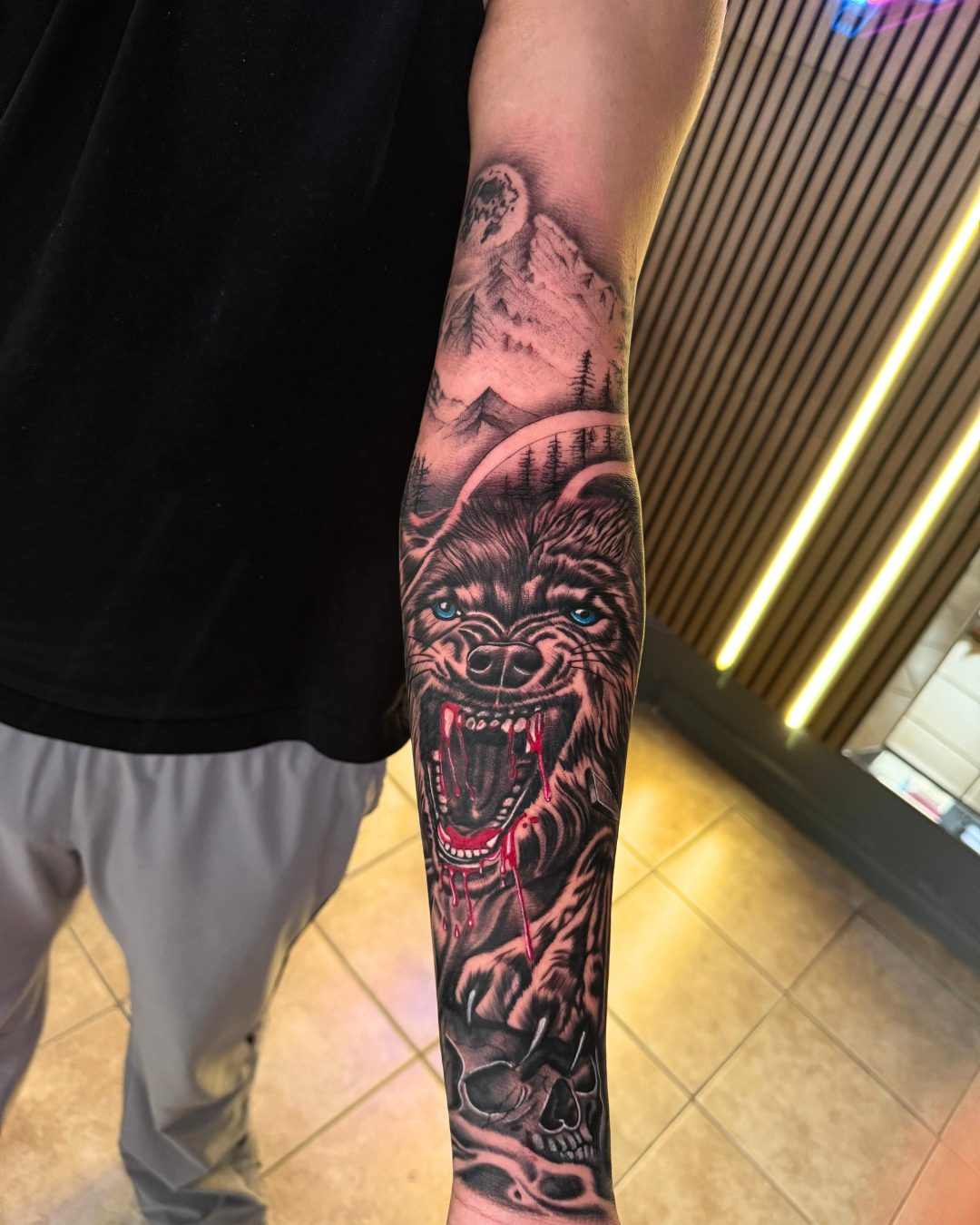 Aggressive black and grey wolf head tattoo with blue eyes, red blood, and a skull, set against a mountain and forest background on the forearm
