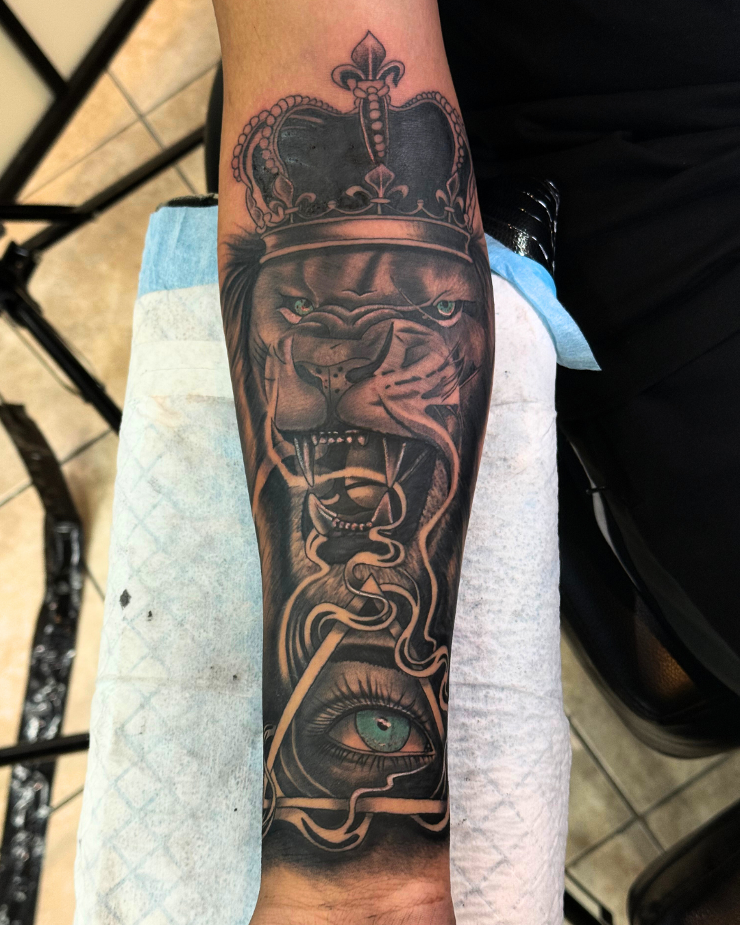 Black and grey lion wearing a crown, above an eye inside a triangle or pyramid on the forearm