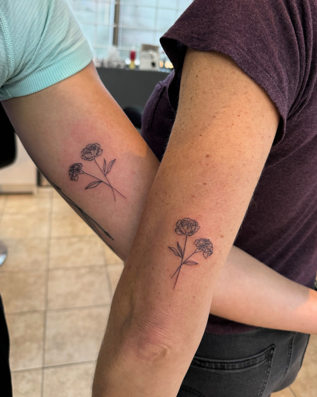 Matching minimalist fine line carnation flower tattoos on the forearms of two people