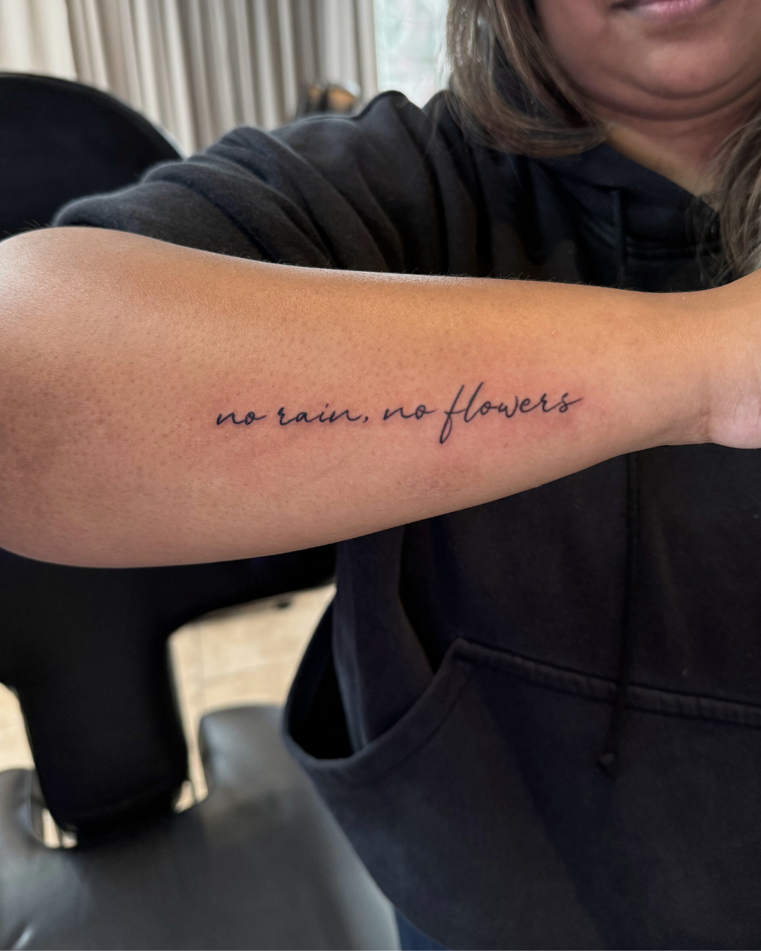 Fine line script tattoo reading no rain, no flowers on the forearm