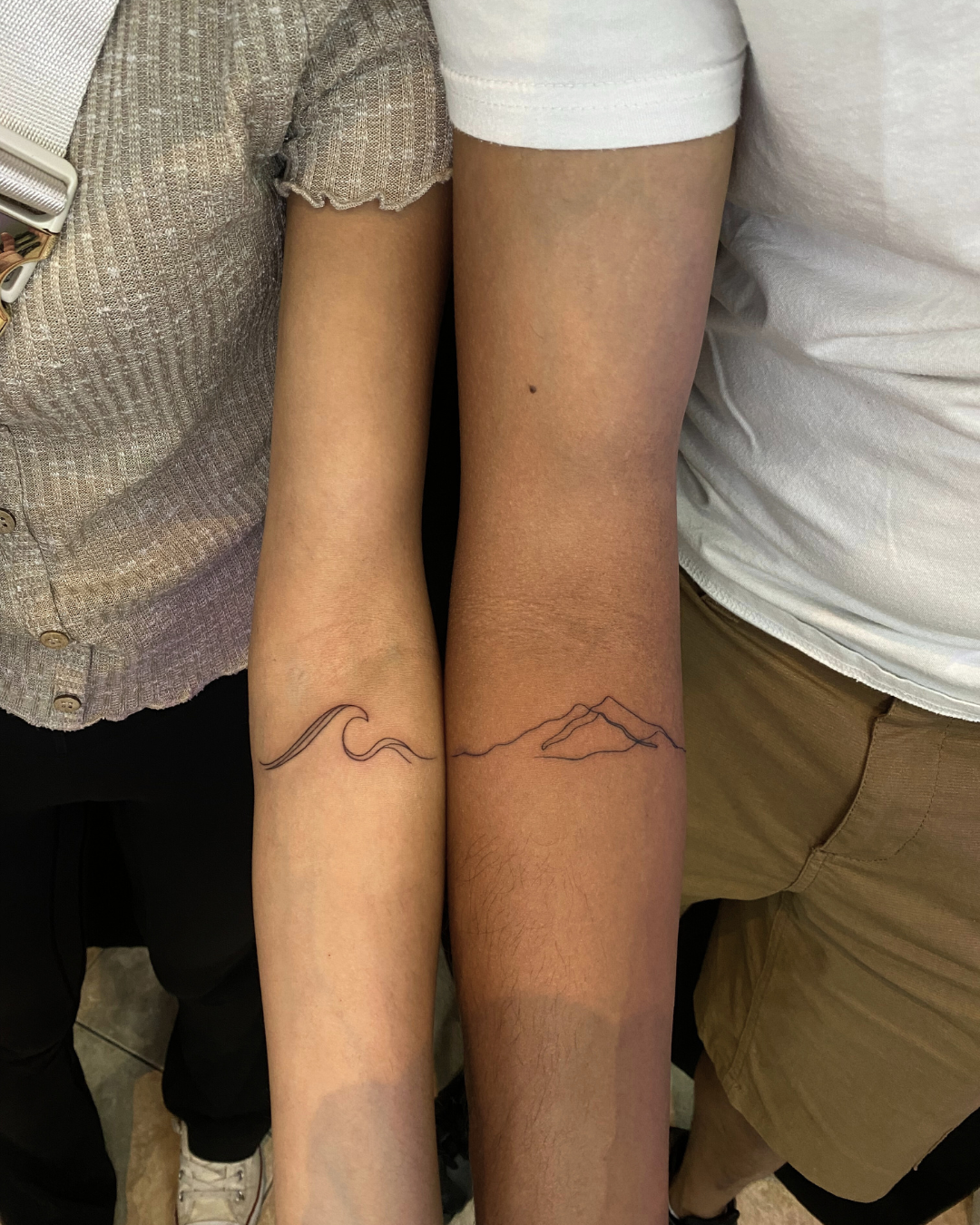 Matching minimalist fine line tattoos of a wave and a mountain on the forearms of two people