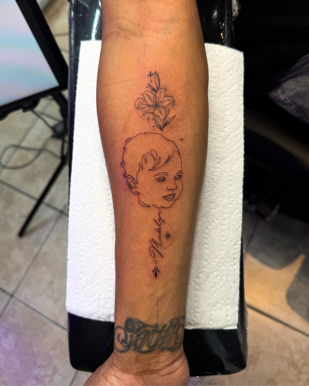 Fine line portrait of a baby with a lily flower above and the name Madelyn below on the forearm
