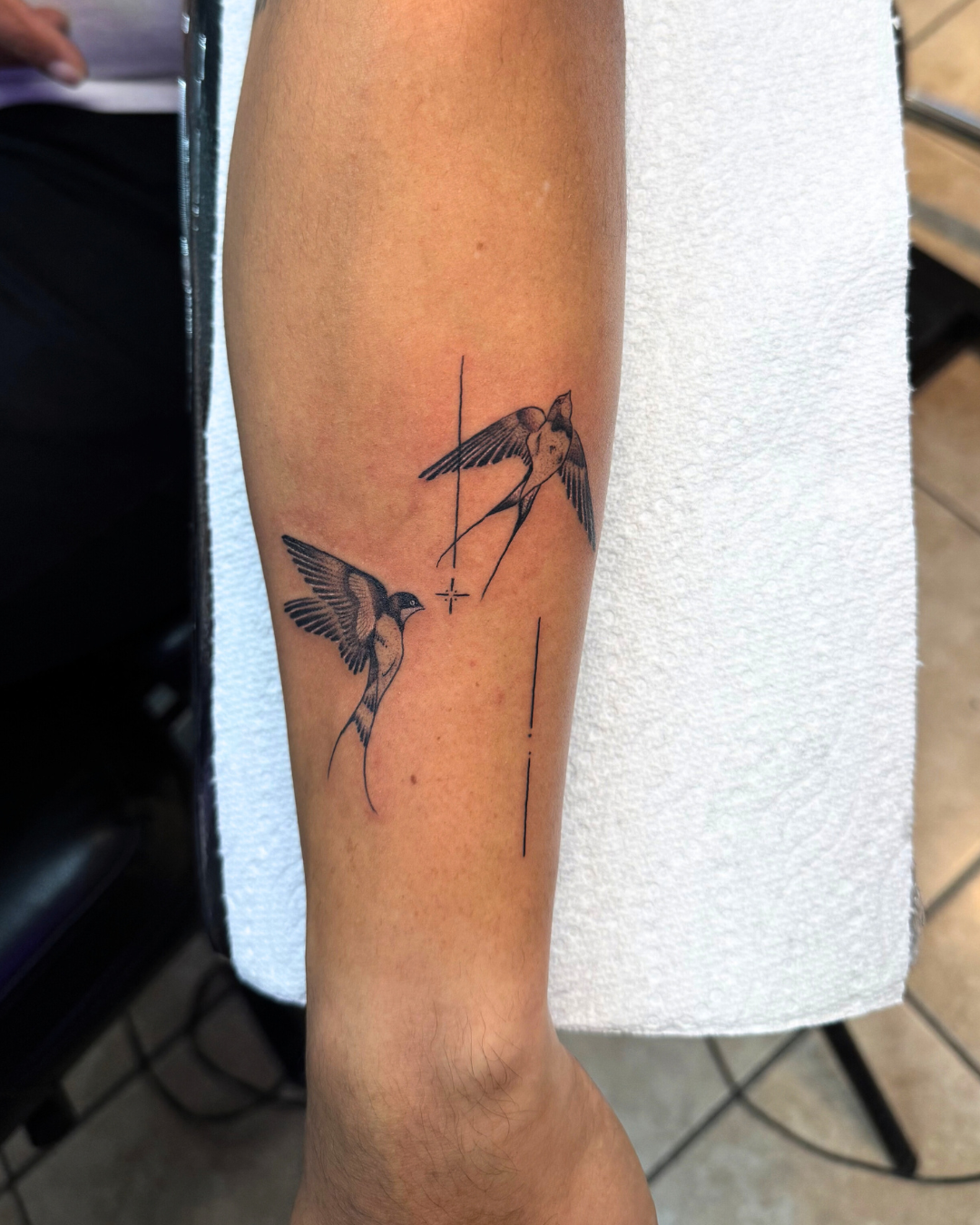 Fine line tattoo of two swallows in flight with minimalist cross and line details on the forearm