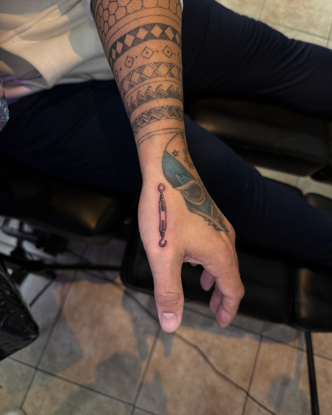 Small fine line tattoo of a turnbuckle or hook on the back of the hand near the thumb