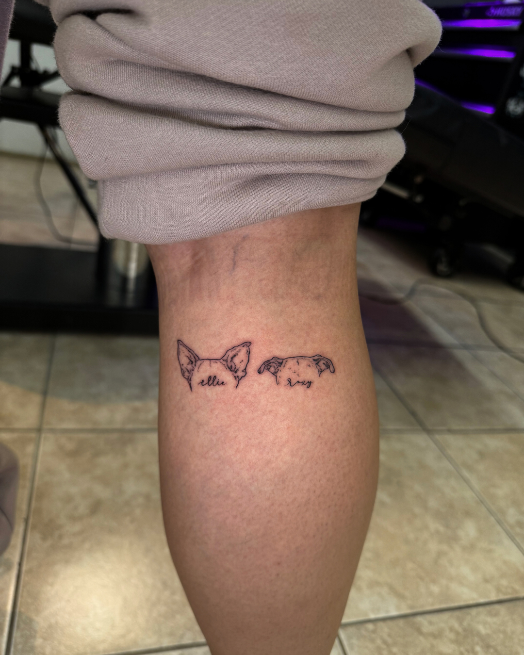 Minimalist fine line tattoos of the tops of two dog heads with the names ellie and roxy on the calf