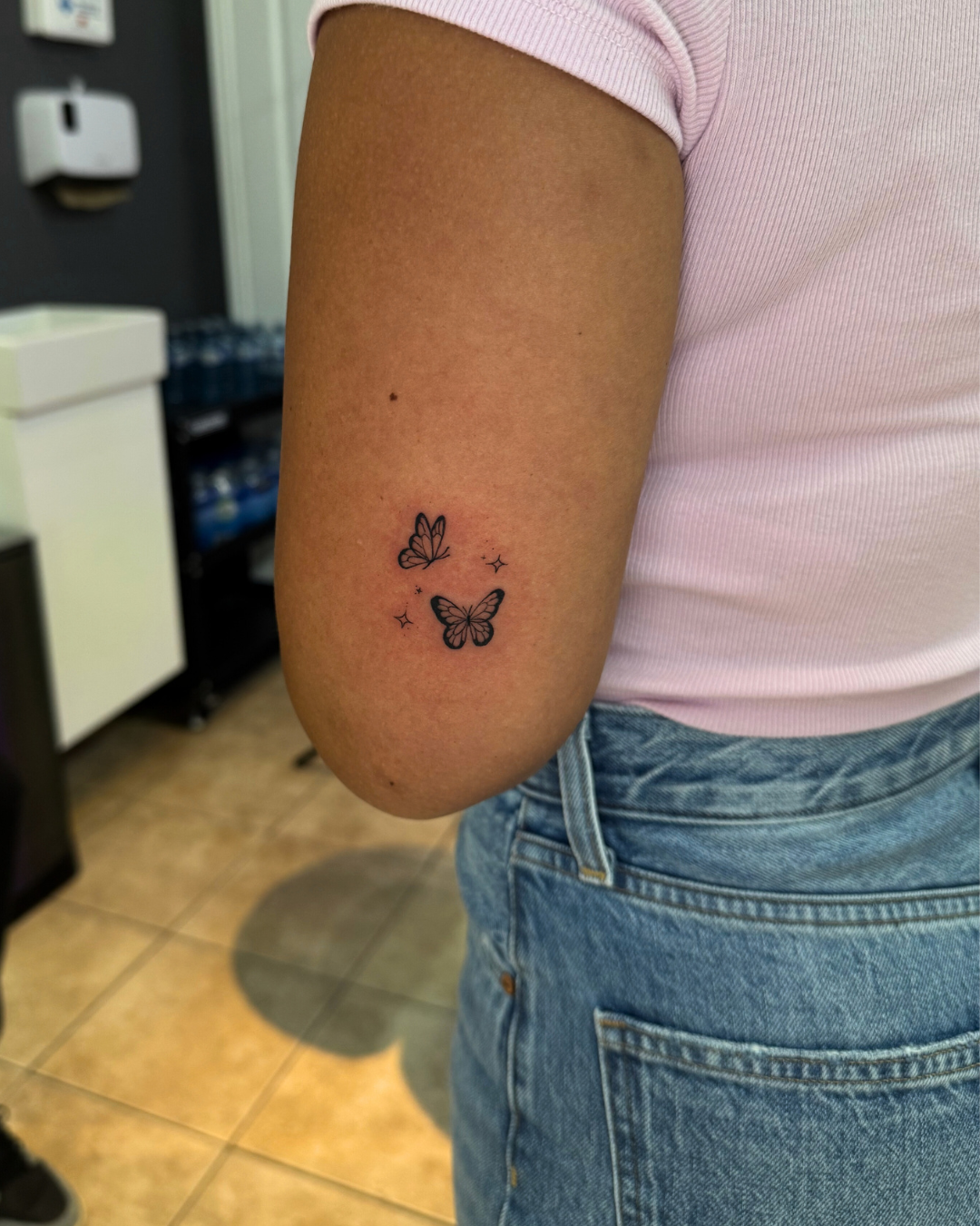 Two small minimalist fine line butterfly tattoos with sparkle details above the elbow