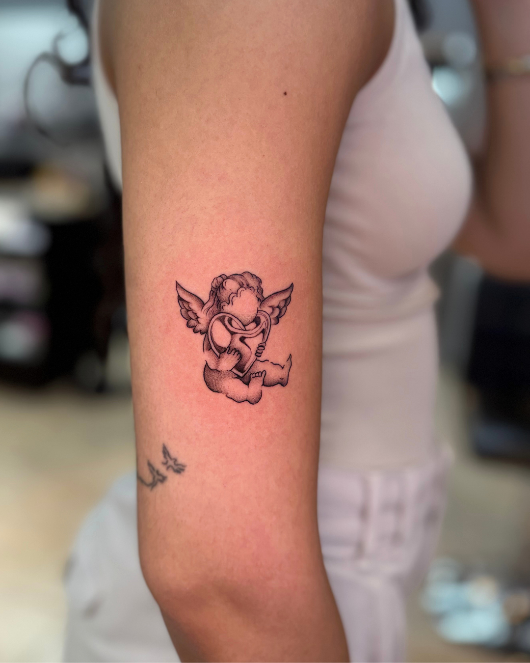 Small fine line cherub angel holding a heart on the upper arm
