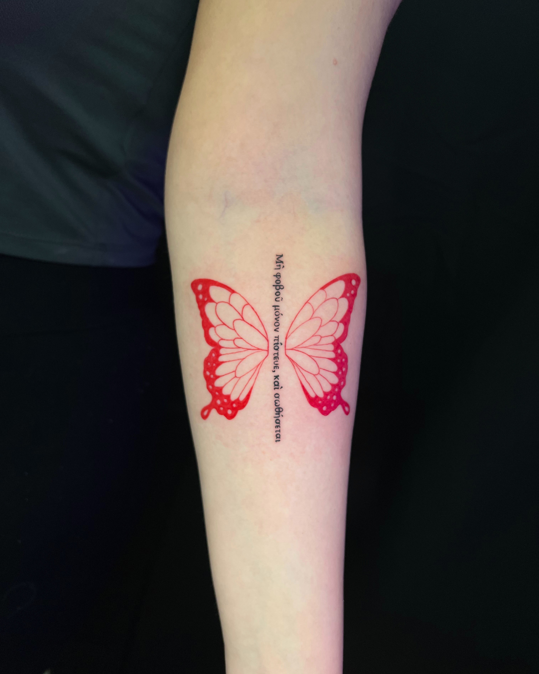 Red ink tattoo of a butterfly with Greek script running vertically through its body on the forearm