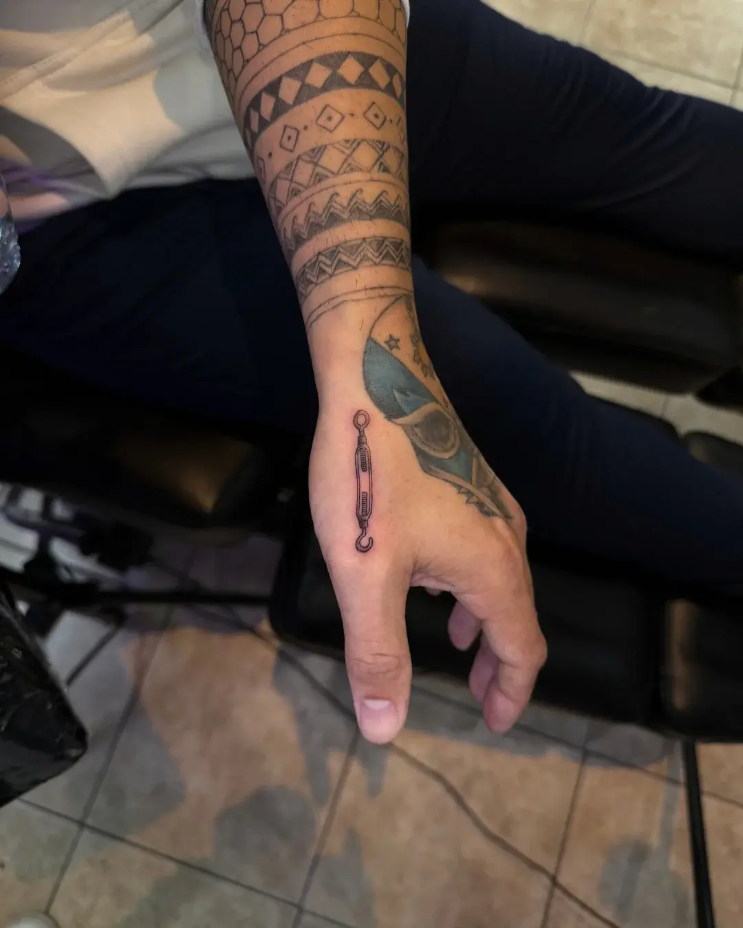Small fine line tattoo of a turnbuckle or hook on the back of the hand near the thumb